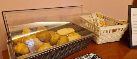 A buffet area with a glass-covered container of pastries and a wicker basket with bread.