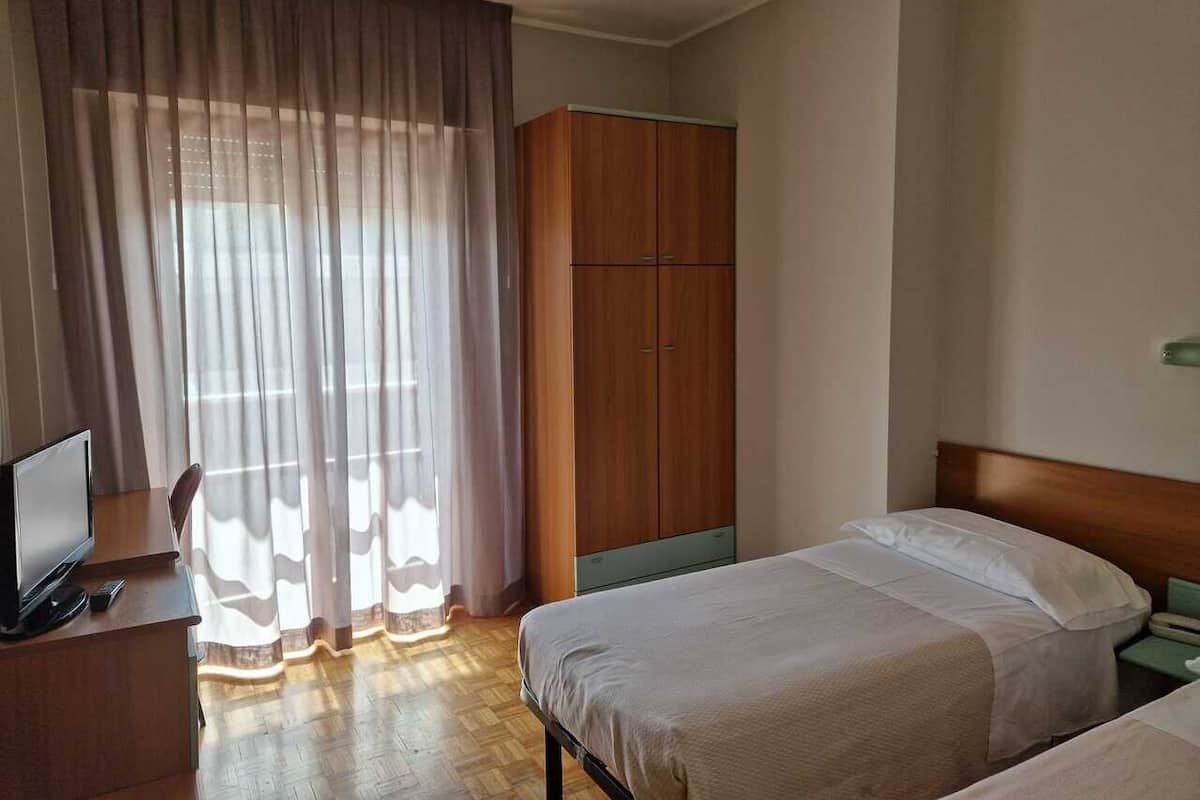 Et hotelværelse med seng, skrivebord med computer, garderobeskab og et vindue med gardiner.