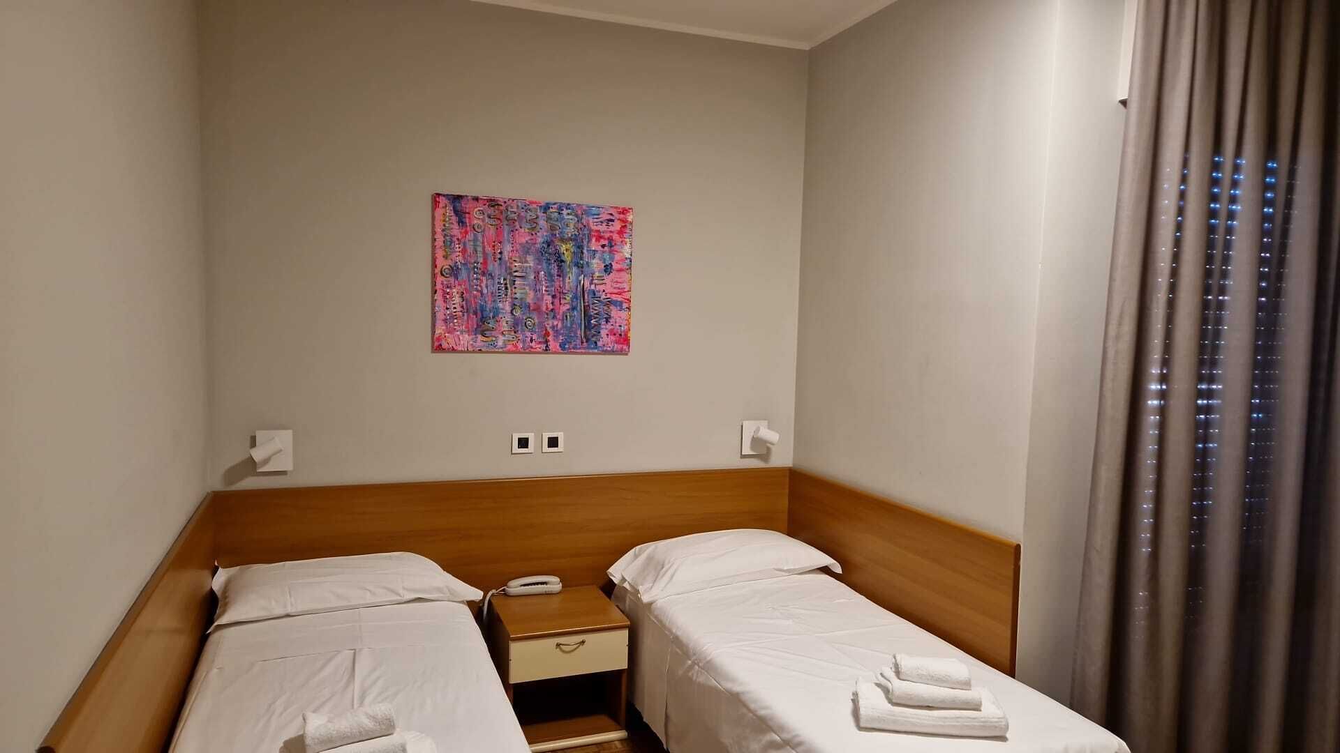 Habitación de hotel con dos camas individuales, cabecero de madera, mesita de noche pequeña, teléfono y un cuadro abstracto colorido en la pared.