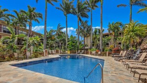 A pool area with a clear blue pool, stone flooring, lounge chairs, palm trees, and a building with balconies. - Prince Kuhio 202, Comfortable Poipu lodging, lanai overlooking the ocean (Poipu)