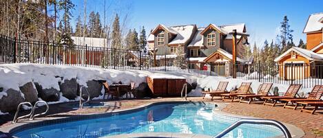 Un espace piscine doté de chaises longues et d’un jacuzzi, entouré de bâtiments et d’un sol recouvert de neige.