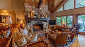 A stone fireplace with a mounted deer head, a cozy seating area with leather sofas, and a large window offering a view of the outdoors. - The Boulders: 5 Suites | Hot Tub | 50mi Views | Pool Table | Sauna | Wet Bar | Dog Friendly! (Boone)
