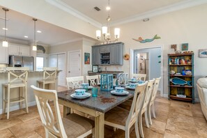 A dining area with a large table set for six, surrounded by white chairs. There are hanging pendant lights above the table, and a blue lantern-style light fixture above the dining table. The room has a neutral color palette with beige walls and a tiled floor.