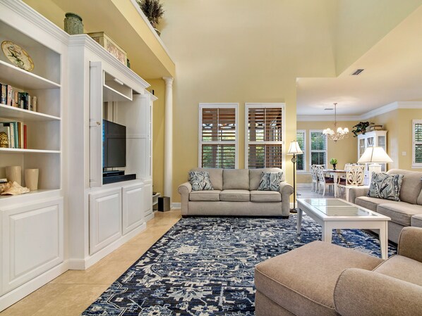 A spacious living room with a large sofa, a coffee table, and a TV. There is a bookshelf and a dining area in the background.