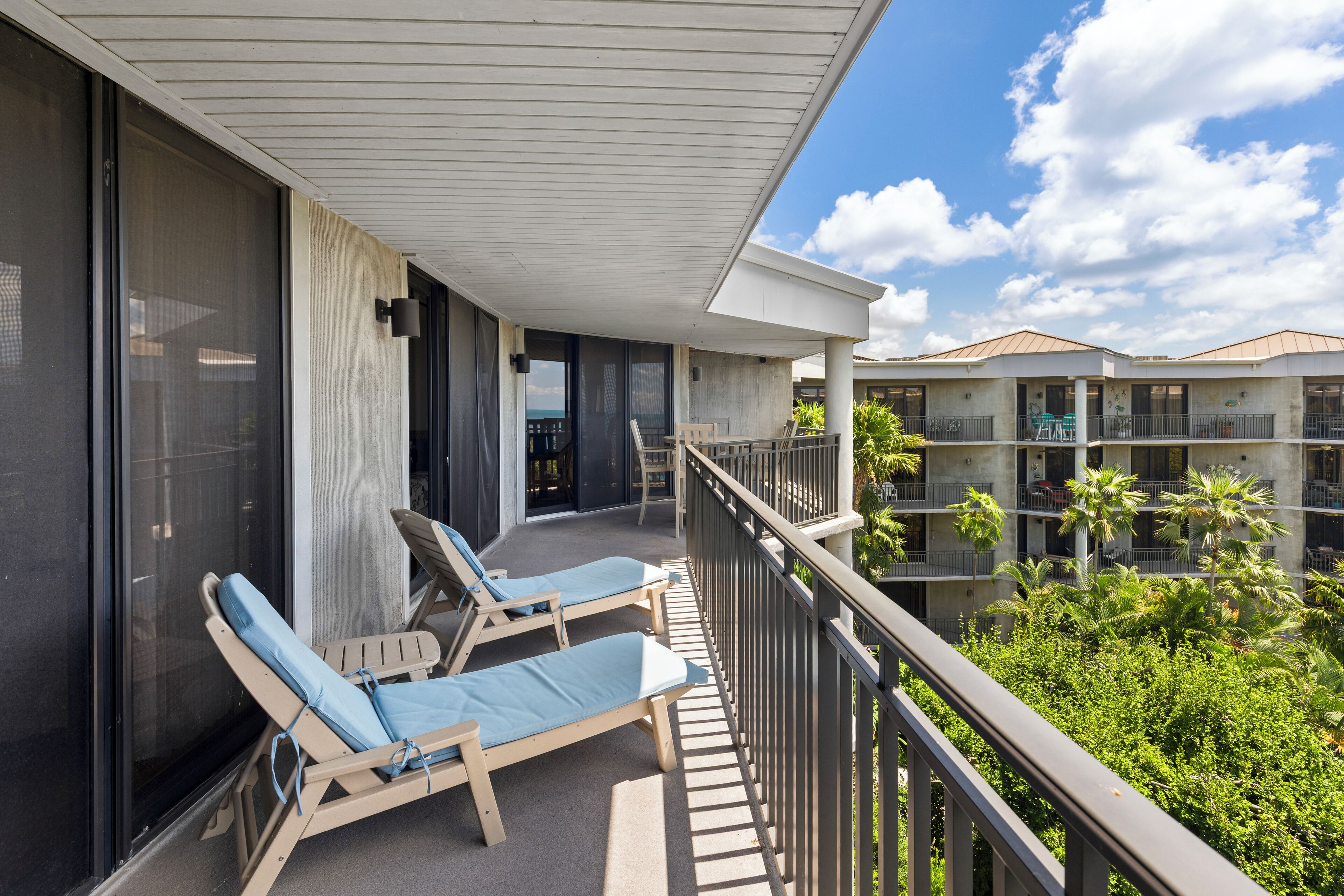 A balcony with two lounge chairs, a view of the ocean, and a building with multiple balconies and palm trees.