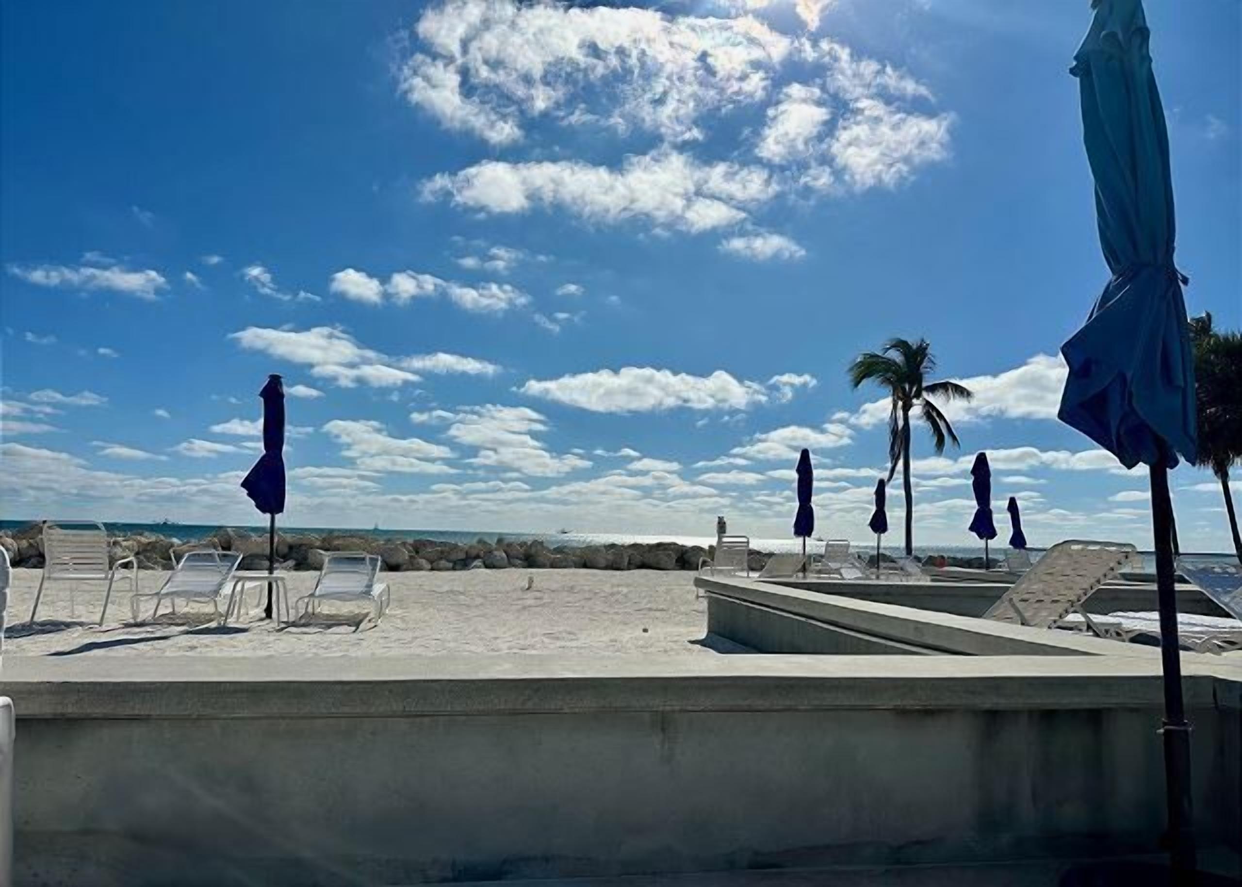 A beachside area with lounge chairs and umbrellas, a concrete pool, and a view of the ocean.