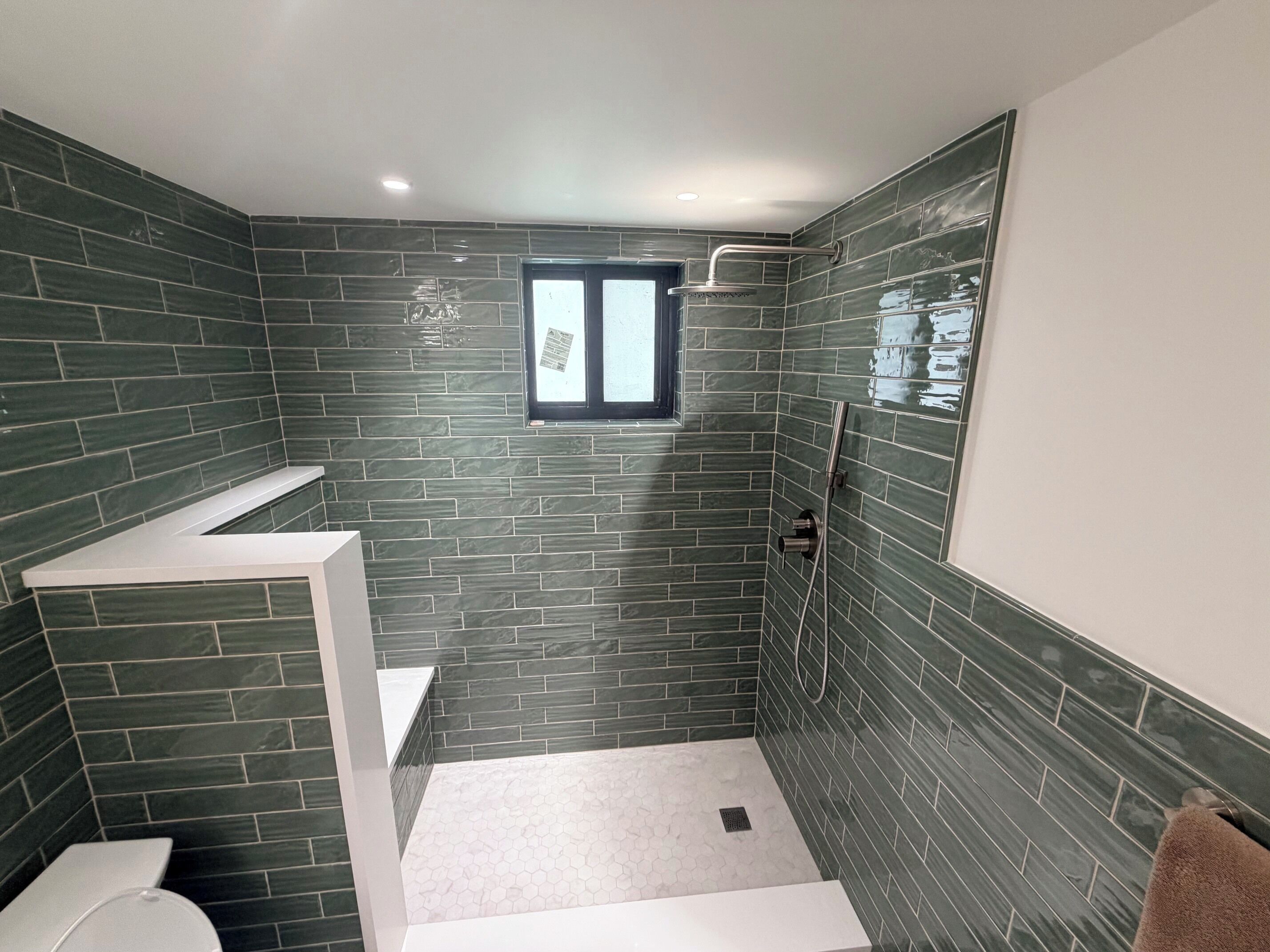 Un baño moderno con azulejos verde oscuro, ducha, inodoro y ventana.
