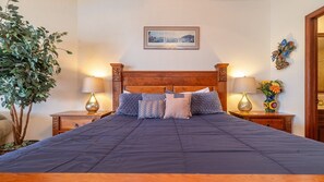 Un lit bien fait, recouvert d’une couverture bleu foncé, est encadré de deux tables de chevet sur lesquelles reposent des lampes et des vases décoratifs. Un tableau encadré est accroché au-dessus du lit, et une plante en pot est visible sur la gauche.