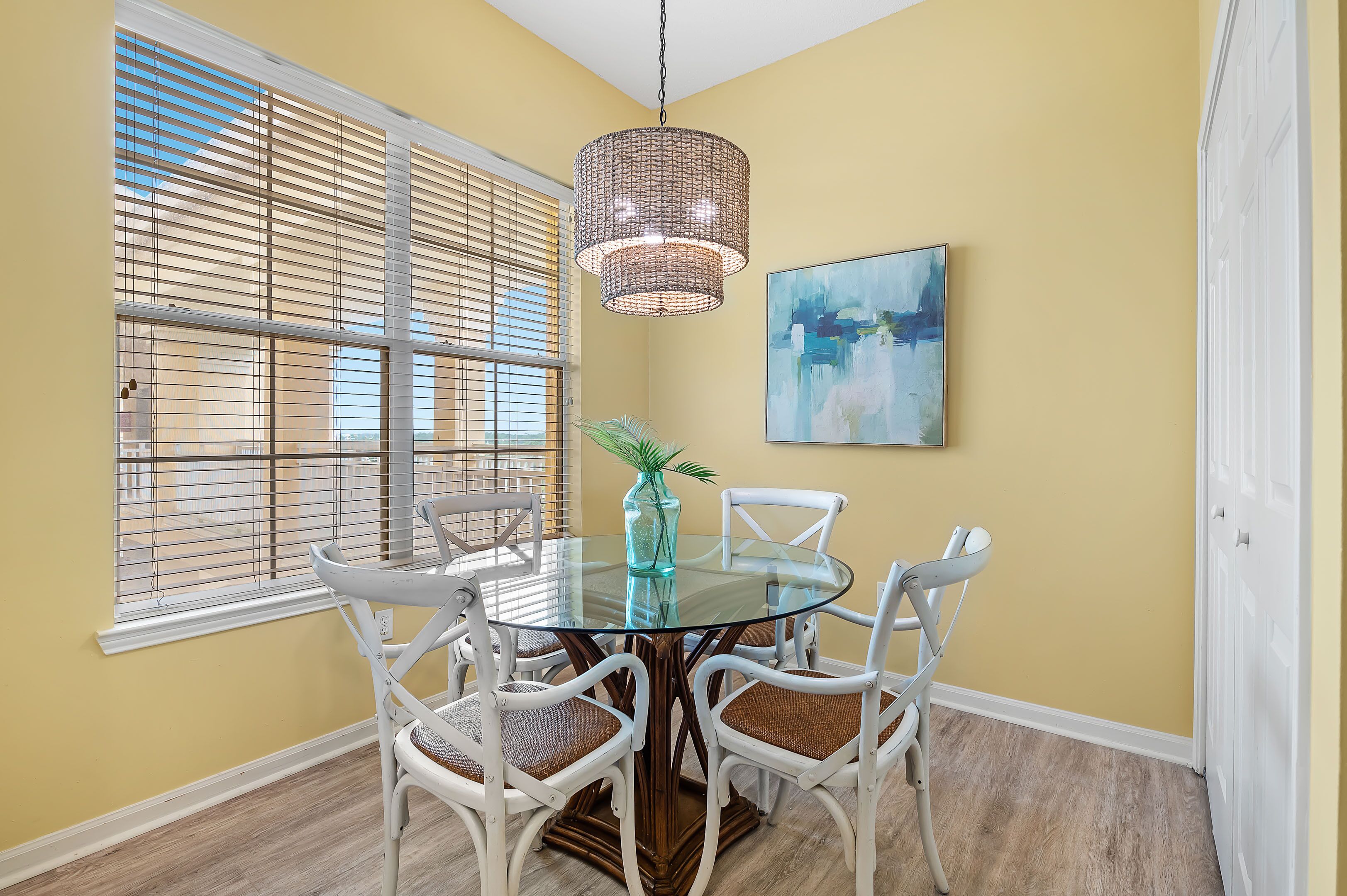 A dining area with a round glass table, white chairs, a hanging light fixture, and a painting on the wall.