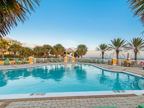 A clear blue pool surrounded by palm trees and a gazebo, with a view of the ocean and a yellow building in the background.