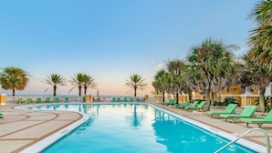 A pool area with a clear view of the ocean, surrounded by palm trees and green lounge chairs.