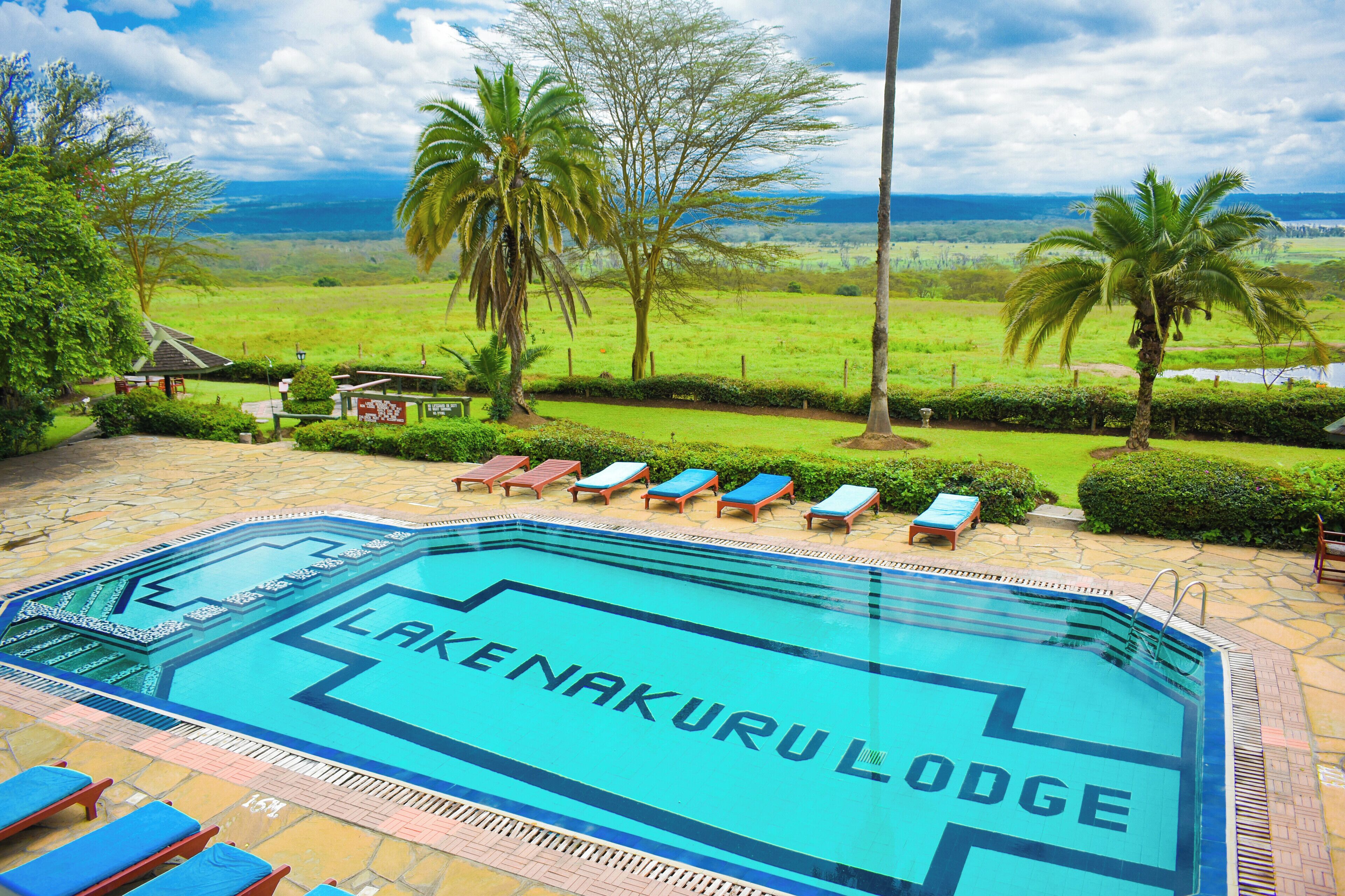 A swimming pool with 'LAKENAKURULodge' written on it, surrounded by lounge chairs and palm trees, with a view of a green landscape.
