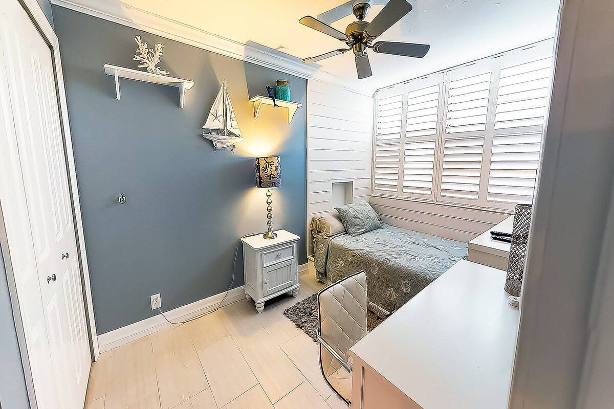 A bedroom with a bed, bedside table, nightstand, wall-mounted shelf with decorative items, and a window with blinds.