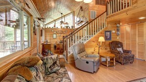 A cozy cabin interior with a wooden floor, a sofa with a bear-themed throw, a recliner, a dining table with chairs, a staircase, and a ceiling fan. - Flat drive- 3BR cabin located between PF and GBurg with hot tub & game room! (Sevierville)