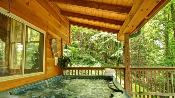 A wooden deck with a hot tub, surrounded by lush greenery. - Flat drive- 3BR cabin located between PF and GBurg with hot tub & game room! (Sevierville)