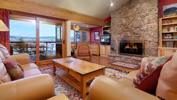 A cozy living room with a stone fireplace, leather sofas, a wooden coffee table, and a large window overlooking a snowy landscape. - JHRL - Snow Ridge #5 - Slopeside Mountain Lodge Decor, close walk to the base (Teton Village)
