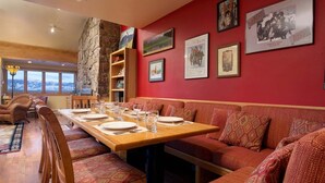 A dining area with a long wooden table set for six, surrounded by cushioned benches. There are framed pictures on the wall, a stone wall feature, and a view through large windows. - JHRL - Snow Ridge #5 - Slopeside Mountain Lodge Decor, close walk to the base (Teton Village)
