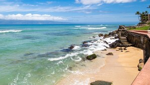Ein Strand mit klarem türkisblauem Wasser, einem sandigen Ufer und einer Steintreppe, die zum Wasser hinabführt.