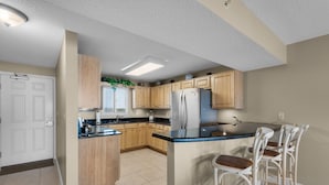 A kitchen with wooden cabinets, a stainless steel refrigerator, and a bar area with stools. - Sand Key 714:  2 BR / 2 BA Gulf Front Condo in Perdido Key, FL  Sleeps 6 (Pensacola)