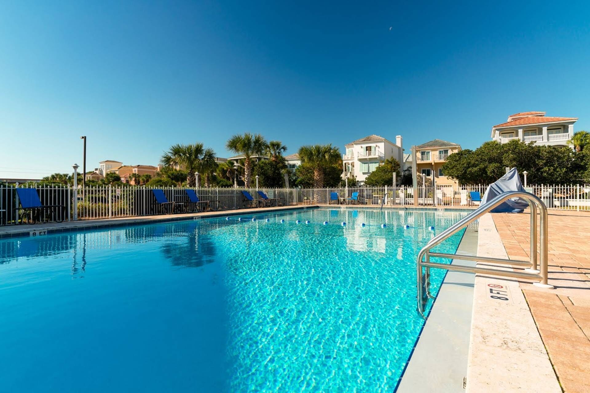 A clear swimming pool with a metal safety railing, surrounded by a white fence and palm trees.