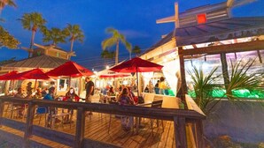 An outdoor dining area with wooden tables and red umbrellas, surrounded by palm trees and a wooden deck. - Hideaway Studio U619 Little Harbor Resort - Tampa Bay (Ruskin)