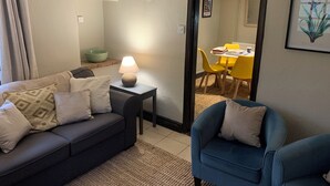 A living room with a dark sofa, a blue armchair, a small table with a lamp, and a dining area with yellow chairs. - Well Cottage in Central Holt Norfolk.  Cosy. Sleeps 4 adults. Free Parking. (Holt, Norfolk)