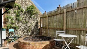 A circular brick fire pit surrounded by white metal chairs and a small round table, set against a stone wall and wooden fence. - Well Cottage in Central Holt Norfolk.  Cosy. Sleeps 4 adults. Free Parking. (Holt, Norfolk)