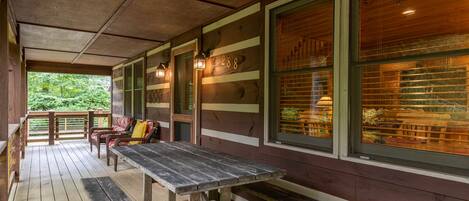 A wooden deck with a picnic table and benches, large windows with blinds, and a view of greenery.