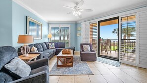 Una sala de estar con ventilador de techo, un conjunto de sofás, una mesa de centro y vistas a un patio exterior por medio de puertas corredizas de vidrio.