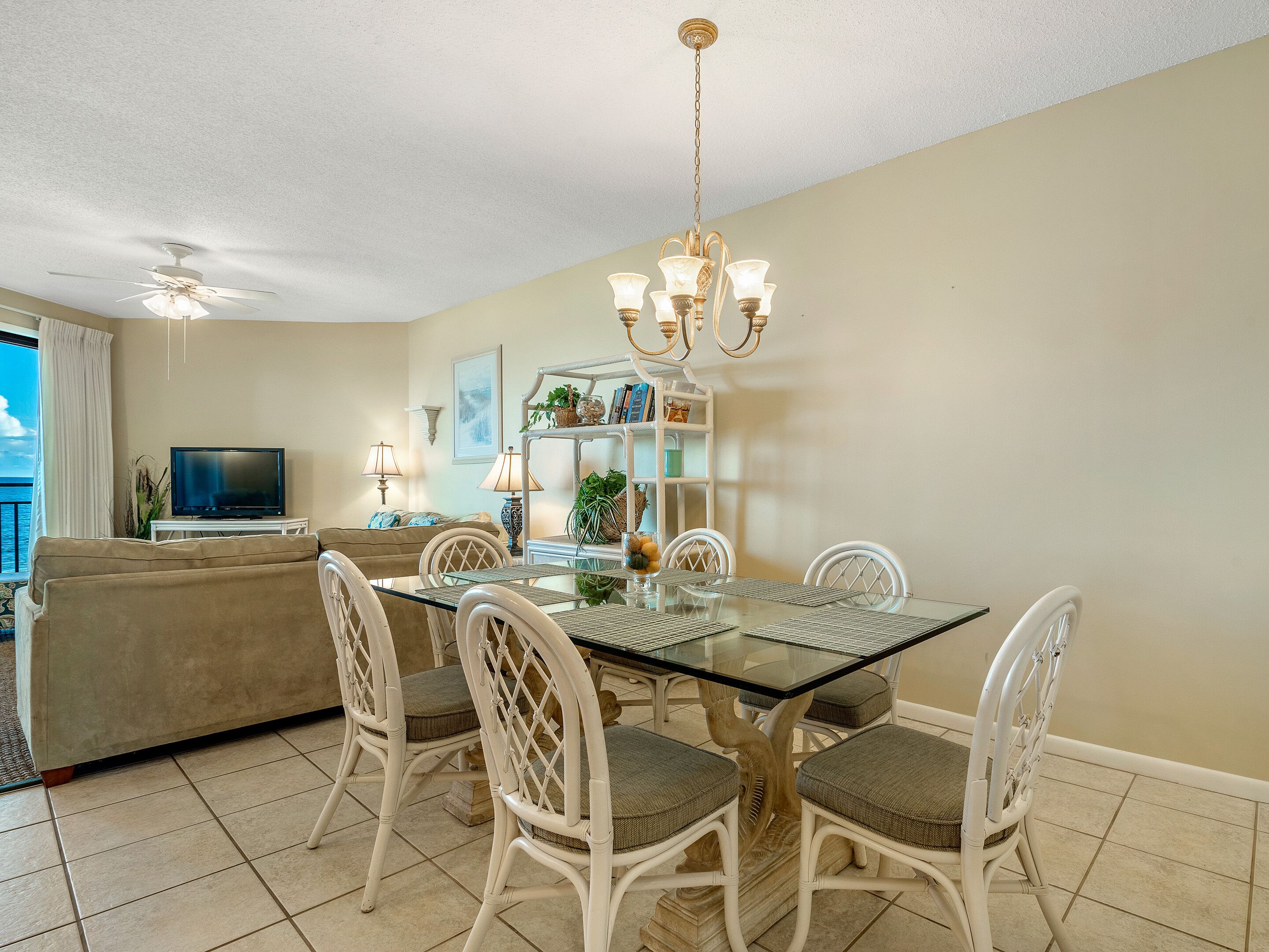 A dining area with a glass table, white chairs, and a chandelier. There is a sofa and a TV in the background.