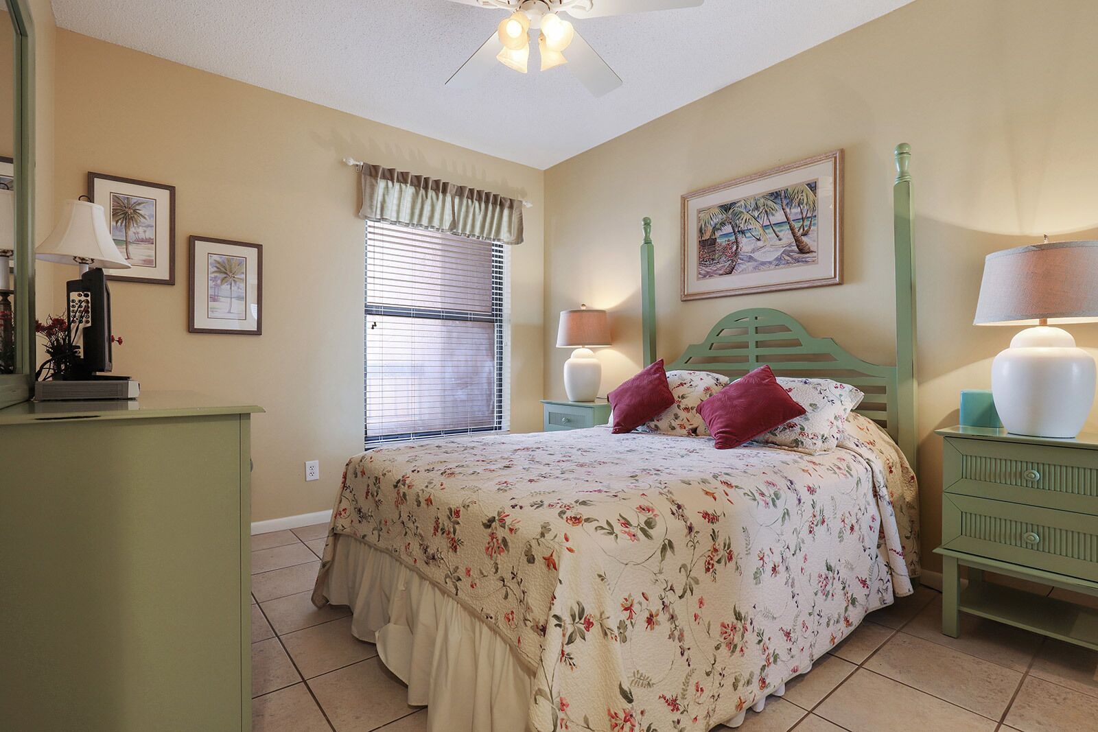 A bedroom with a bed, two bedside tables, a nightstand, a window with blinds, and a ceiling fan.