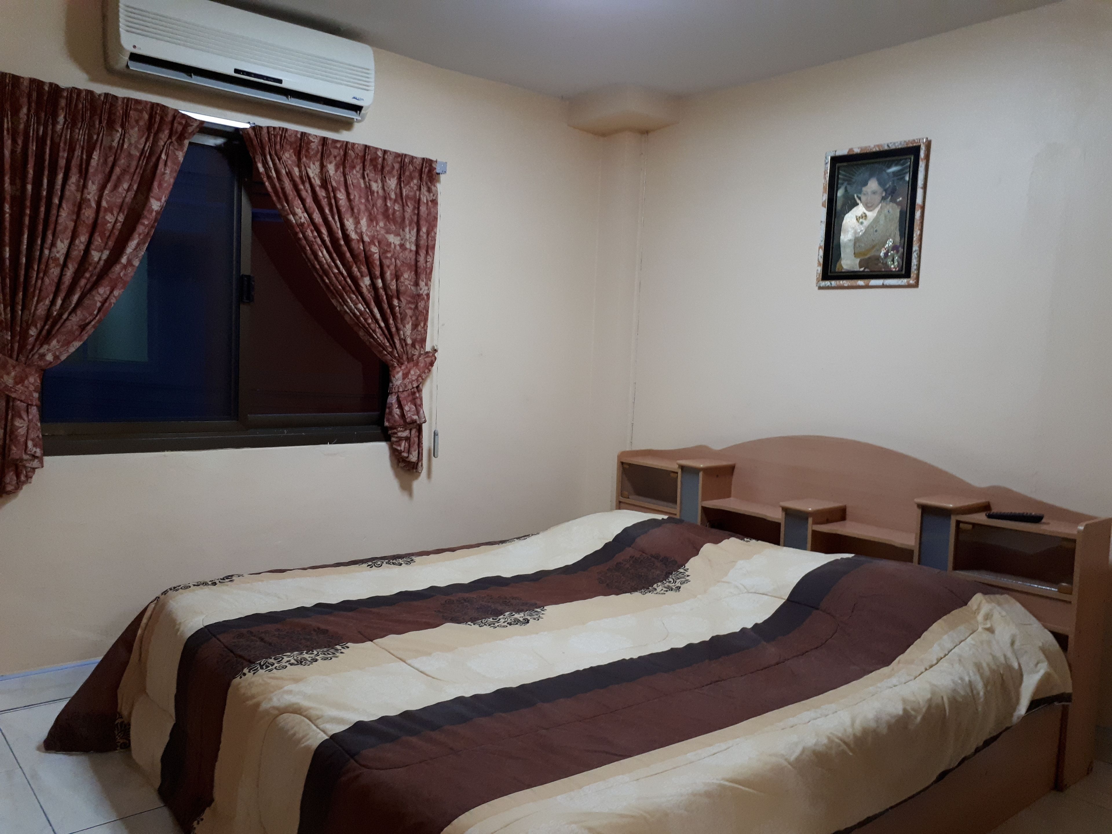 Un dormitorio con cama, ventana con cortinas, aire acondicionado, un cuadro en la pared y cabecera de madera.
