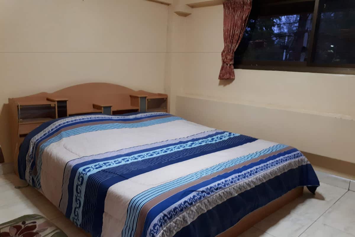 A bedroom with a bed, a wooden headboard, a window with curtains, and a tiled floor.