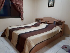 A single bed with a wooden headboard and side tables, a window with curtains, and a framed picture on the wall. - Urs Treff Guesthouse (Cha-am)