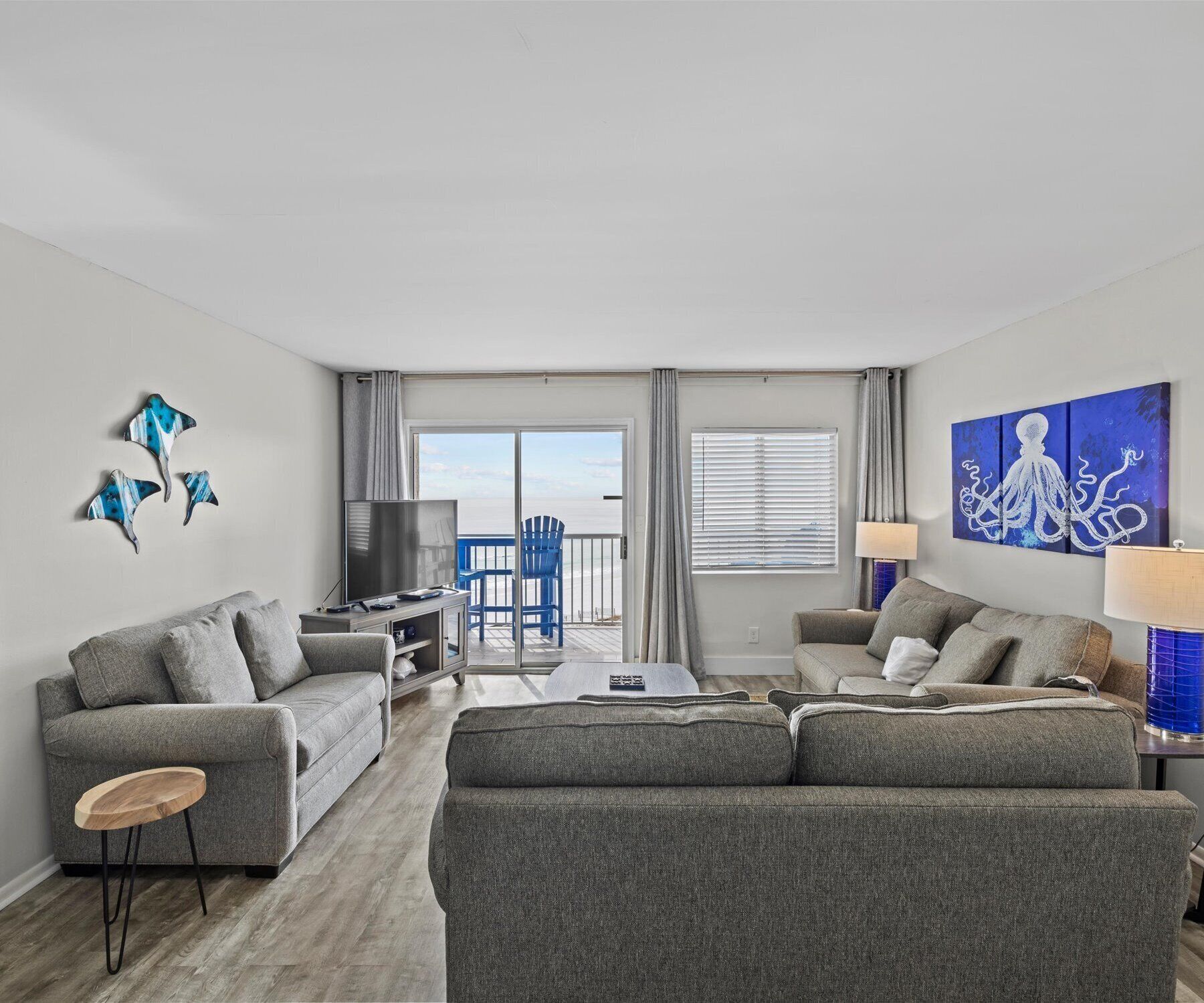 A modern living room with a grey sofa, a coffee table, and a TV. There is a balcony with a blue chair and a view of the sea.