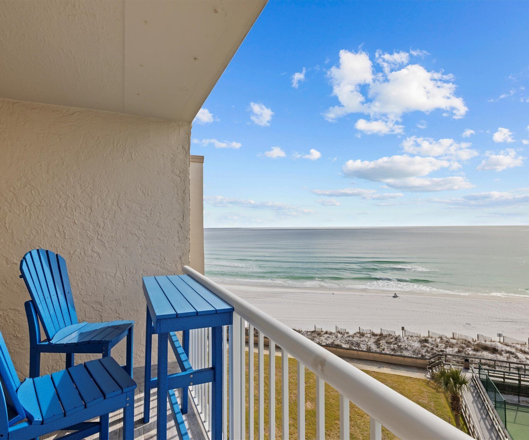 A balcony with a view of the beach, featuring two blue Adirondack chairs and a white railing.