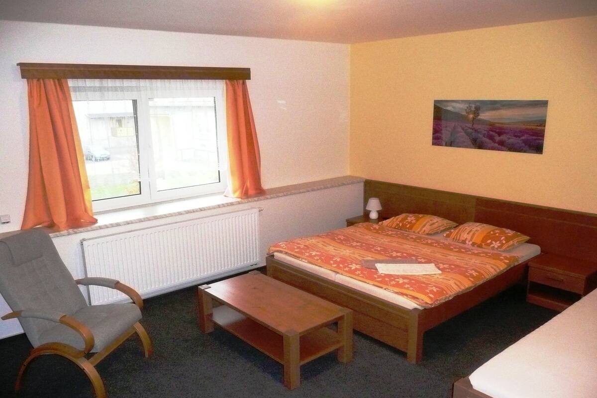 A bedroom with a bed, a chair, a small table, a radiator, and a window with curtains.