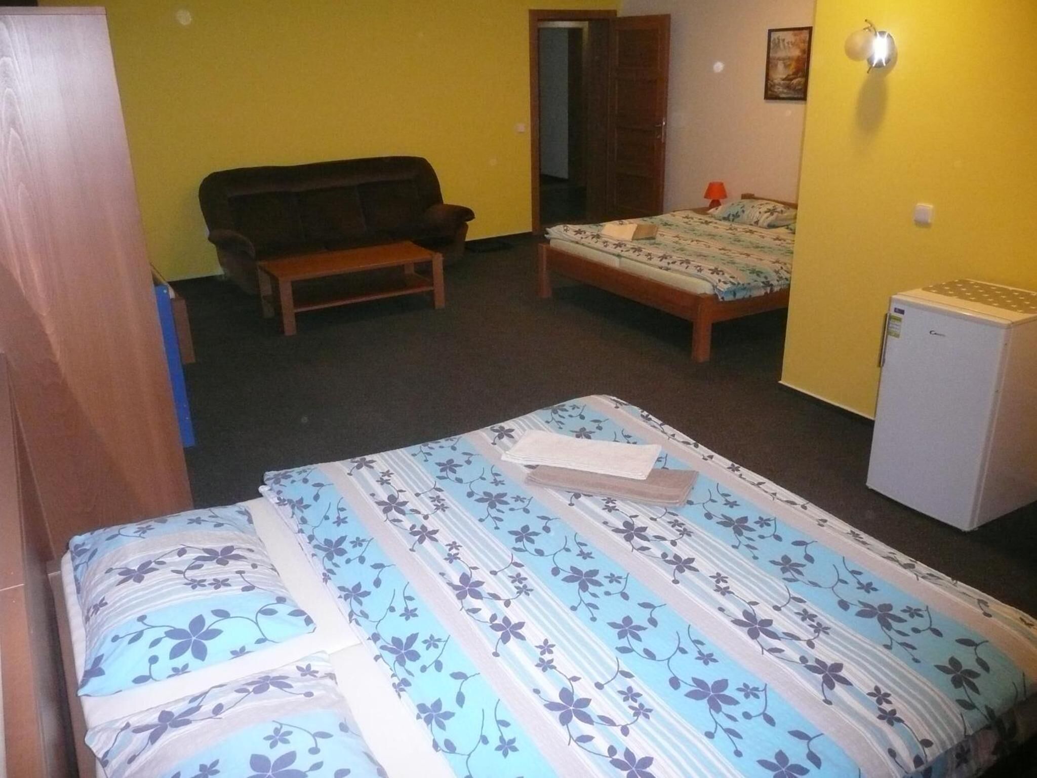 Quarto pequeno e compacto com uma cama, um banco, um refrigerador e uma porta que dá para outro espaço.