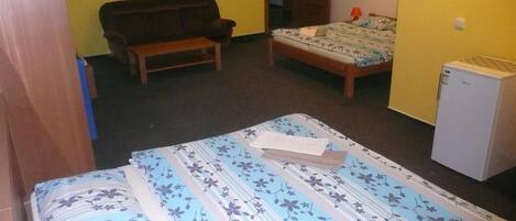 Quarto pequeno e compacto com uma cama, um banco, um refrigerador e uma porta que dá para outro espaço.