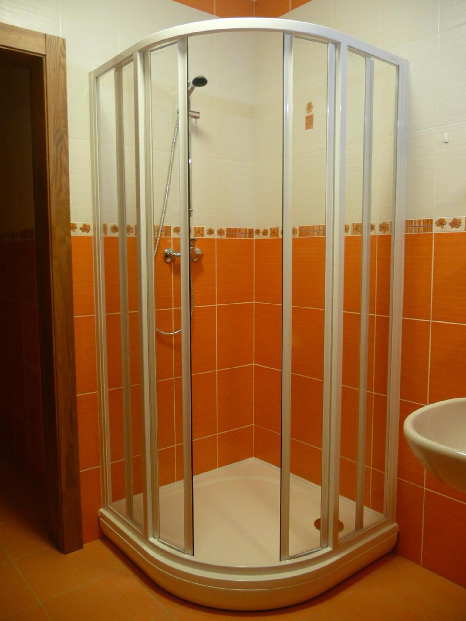 A corner shower with a glass enclosure, orange tiled walls, and a white sink.