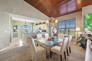 Uma sala de jantar espaçosa com uma mesa de vidro rodeada por cadeiras acolchoadas, um candelabro e vista para a cozinha e sala de estar.