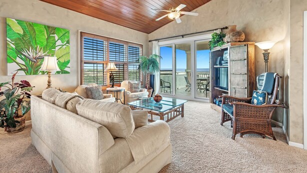 A spacious living room with a high ceiling, a ceiling fan, a large window with blinds, a glass coffee table, a beige sofa, wicker chairs, and a potted plant.