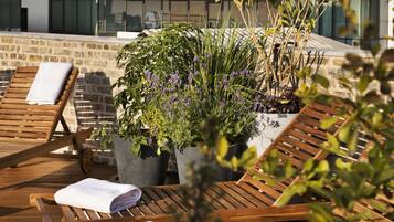 A rooftop terrace with wooden lounge chairs, potted plants, and a stone wall.
