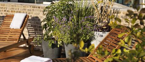 A rooftop terrace with wooden lounge chairs, potted plants, and a stone wall.