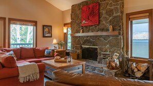A cozy living room with a stone fireplace, a red painting, and a large window overlooking a body of water. - JHRL - Slope-side Rendezvous C4 condo, steps from Hot tub, sleeps 9 (Teton Village)