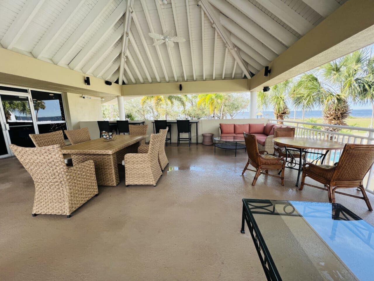 Un patio techado al aire libre con mobiliario de mimbre, una mesa de vidrio y vista al océano.
