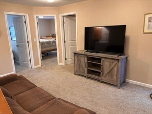 A living room with a brown sofa, a TV on a wooden stand, and a door leading to a bedroom.