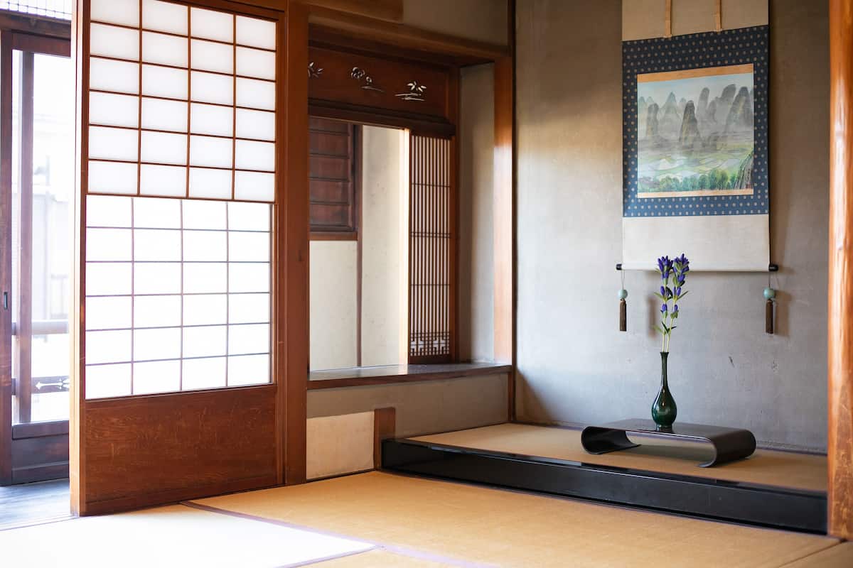 Ein traditioneller japanischer Raum mit Tatami-Boden, einer Shoji-Schiebetür, einer Wandmalerei und einer Vase mit Blumen.
