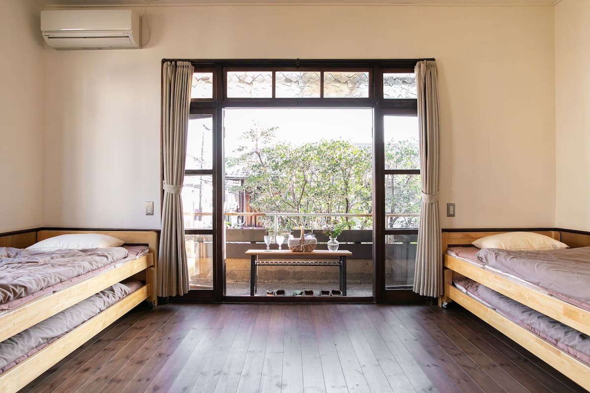 Ein Zimmer mit zwei Holzbettgestellen, einem Holztisch mit Korb und Gläsern sowie einer großen Schiebeglasdoor zur Außenbalkon.