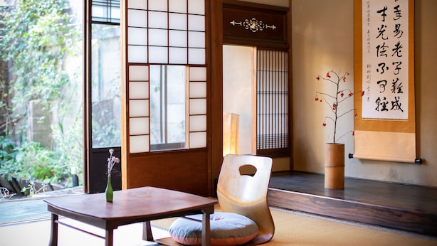 A traditional Japanese room with tatami flooring, a low table, a cushion, a wooden chair, and a sliding door with a view of greenery outside. - Higurashi-Sou (Kyoto)